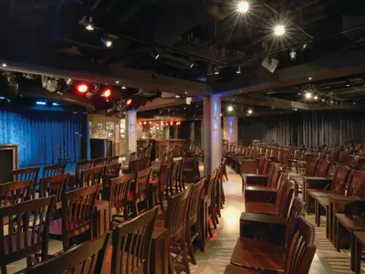 Der Headliners Comedy Club der Norwegian Breakaway. Bühne mit Mikrofon, blaue Vorhänge, viele Holzstühle und eine Bar im Hintergrund bei gedämpftem Licht.