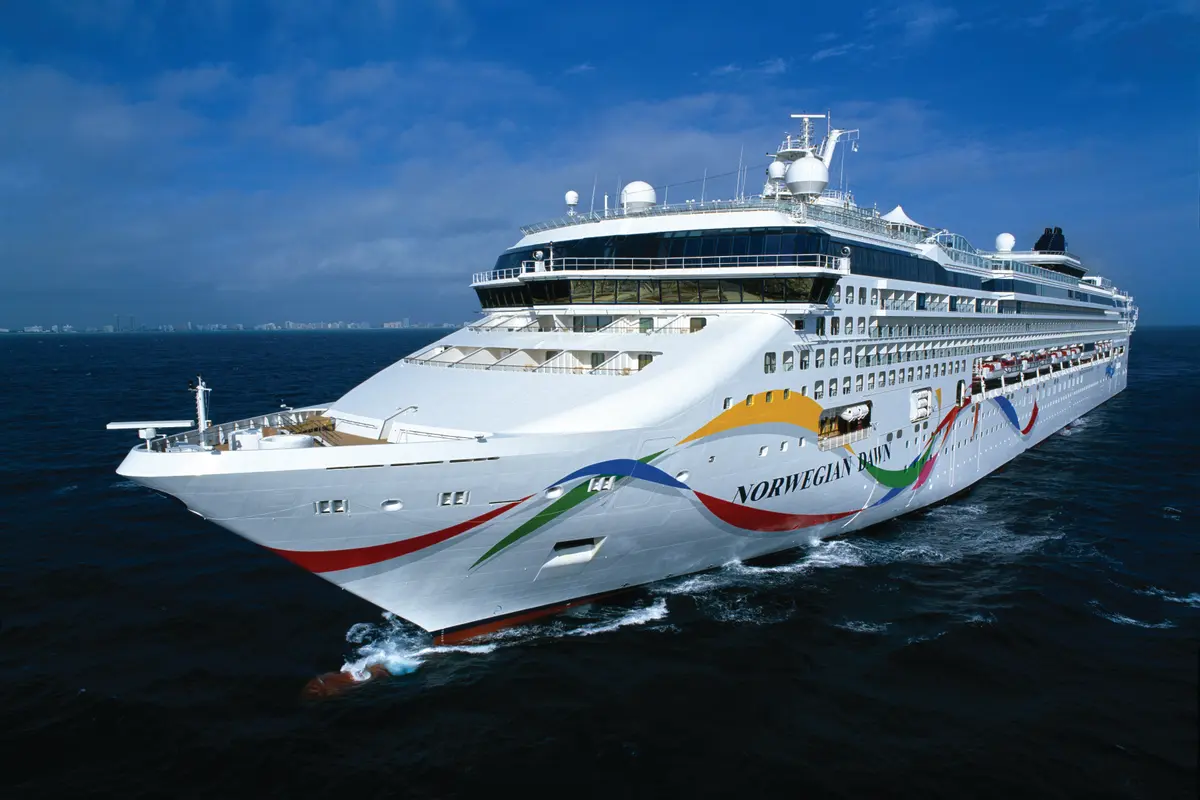 Die Norwegian Dawn auf offener See, der Bug mit farbenfroher Regenbogen-Rumpfkunst. Im Hintergrund eine weite Stadt-Skyline unter blauem Himmel mit Wolkenformationen.
