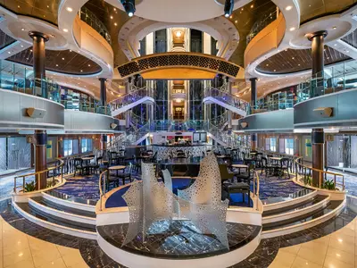 Das mehrstöckige Atrium an Bord der Norwegian Dawn, mit einer zentralen, geschwungenen Bar, umrahmt von symmetrischen Grand Staircases. Moderne Skulpturen im Vordergrund, Glas- und Lichtinstallationen. Der Bereich ist mit Tischen und Stühlen ausgestattet.