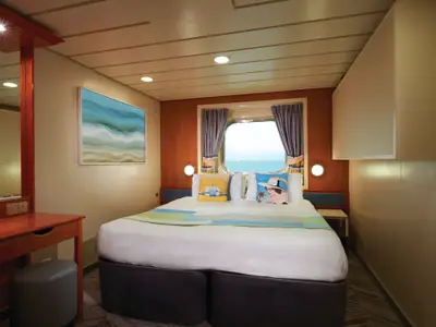 Moderne, helle Aussenkabine an Bord der Norwegian Dawn. Ein Doppelbett mit farbigen Kissen steht vor einem Fenster mit direktem Meerblick. Links ein Schminktisch, rechts ein Wandschrank. Die Kabine ist leer.
