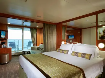 Balkonkabine an Bord der Norwegian Dawn. Das Doppelbett mit weißer Bettwäsche ist im Vordergrund. Links ein Sitzbereich, TV, und ein Balkon mit Meerblick. Helle Holzeinrichtung.