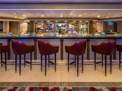 Dunkelbraune Leder-Barhocker stehen an einer beleuchteten Bar der Norwegian Dawn. Im Hintergrund Regale mit Spirituosen, Bildschirme und Spielautomaten einer Casino Bar.