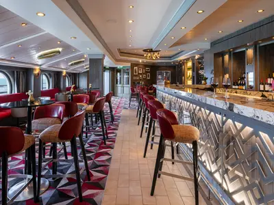 Die elegante Weinbar 'The Cellars' an Bord der Norwegian Dawn. Langer Marmortresen mit Weinflaschen und Gläsern, rote Barhocker und gemütliche Sitzecken. Runde Fenster bieten Meerblick, während ein geometrischer Teppich und warme Beleuchtung das Ambiente prägen.