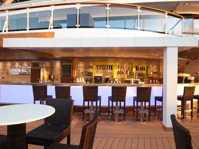 Die moderne Topsider Bar an Bord der Norwegian Dawn auf dem Oberdeck zeigt eine beleuchtete Theke, gut gefuellte Regale mit Flaschen und Barhocker. Einladende Atmosphaere bei Tageslicht.