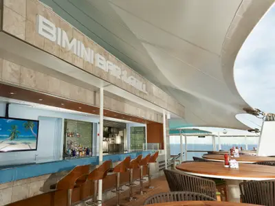 Die Bimini Bar & Grill auf der Norwegian Dawn. Eine Aussenbar mit Tresen, Hockern und TV. Sitzbereich mit Tischen und Stühlen unter einem Sonnensegel, mit Blick auf das offene Meer.