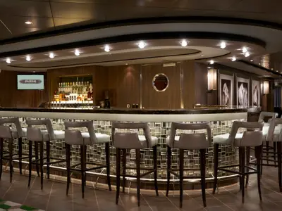 Elegante Sugarcane Bar an Bord der Norwegian Dawn mit beleuchtetem Tresen, grauen Barhockern und einer Spirituosenwand. Holzvertäfelung und indirekte Beleuchtung schaffen eine moderne Atmosphäre. Der Raum ist leer.