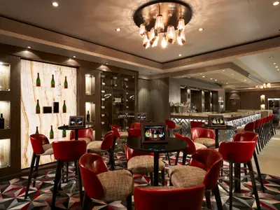Elegante Weinbar an Bord der Norwegian Dawn. Rote Stühle, Tische mit Tablets, beleuchtete Marmorwand mit Weinflaschen und Weinkeller. Einladende Atmosphäre.
