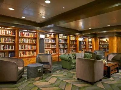 Eine einladende Bibliothek an Bord der Norwegian Dawn, gefuellt mit Buechern, bequemen Sesseln und einer gruenen Couch. Stimmungsvolle Beleuchtung schafft eine ruhige Atmosphaere.