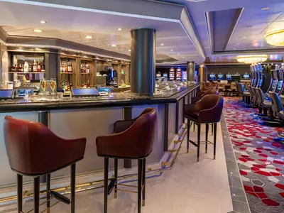 Ein luxuriöses Casino an Bord der Norwegian Dawn mit einer langen Marmorbar, Spielautomaten und braunen Ledersesseln. Roter und grauer Teppich. Warme Beleuchtung.