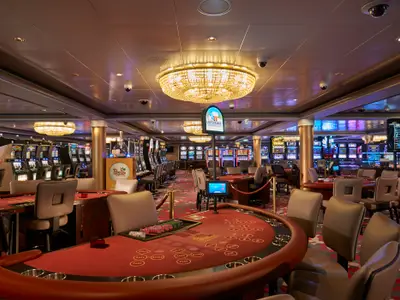 Weitwinkelaufnahme des Casinos an Bord der Norwegian Dawn. Zahlreiche Spielautomaten und rote Spieltische sind unter funkelnden Kronleuchtern in einem modernen Raum zu sehen.
