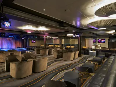 Innenansicht der modernen Club-Lounge an Bord der Norwegian Dawn mit bequemen Sesseln, Sofas und einer Bühne mit Vorhängen und farbigen Scheinwerfern. Elegante Beleuchtung und Teppichboden.