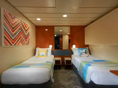 Innenansicht einer hellen Kabine an Bord der Norwegian Dawn mit zwei Einzelbetten, bunter Bettwäsche und einem modernen Kunstwerk. In der Mitte ein Spiegel.