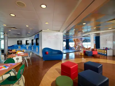 Detailaufnahme des Kinderbereichs Splash Academy an Bord der Norwegian Dawn. Bunte Tische, Stühle, ein blauer Sessel und Wandbilder mit Meeresthema sind zu sehen. Der Raum ist hell beleuchtet und hat runde Fenster.