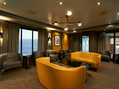 Eine stilvolle Lounge an Bord der Norwegian Dawn mit dunklen Ledersesseln und senffarbenen Ledersofas, die um einen Couchtisch gruppiert sind. Große Fenster bieten einen weiten Blick auf das offene Meer. Warme Beleuchtung und Holzwände schaffen eine gemütliche Atmosphäre.