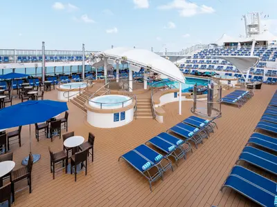 Blick auf das obere Pooldeck der Norwegian Dawn mit einem großen Pool, zwei Whirlpools und Reihen von blauen Liegestühlen. Eine Bar und Sitzbereiche sind ebenfalls zu sehen.