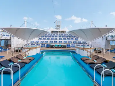 Weitwinkelaufnahme des sonnigen Pooldecks der Norwegian Dawn. Ein langer Pool, umgeben von Liegestühlen, zwei Whirlpools und gestuften Sitzreihen. Blauer Himmel.