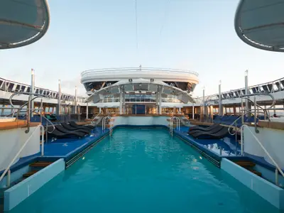 Blick auf den Hauptpool der Norwegian Dawn. Ein türkisfarbener Pool mit schwarzen Sonnenliegen auf blauem Deck. Im Hintergrund die weiße Schiffssuperstruktur unter klarem Himmel. Entspannte Tagesstimmung.