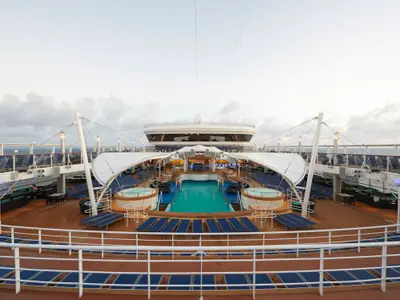 Ein Weitwinkelblick auf das obere Pooldeck der Norwegian Dawn. In der Mitte befinden sich ein großer Pool und zwei Whirlpools, umgeben von zahlreichen blauen Liegestühlen und einem Holzdeck. Im Hintergrund ist die Brücke des Schiffes zu sehen. Der Himmel ist bewölkt und hell.
