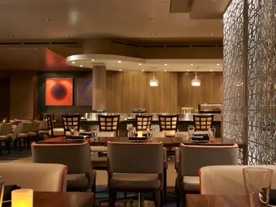 Das Restaurant Bamboo an Bord der Norwegian Dawn zeigt moderne Tische, Stühle, Holzwände, abstrakte Kunst und warme Beleuchtung. Eine dekorative Trennwand ist rechts sichtbar.