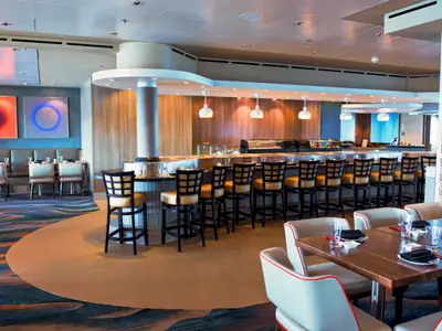 Ein weitläufiges Spezialitätenrestaurant an Bord der Norwegian Dawn, mit einer langen Sushi-Bar, hohen Hockern und Tischen mit bequemen Stühlen. Moderne Kunst und Holzwände schmücken den Raum.