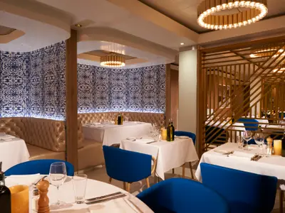 Elegantes Restaurant an Bord der Norwegian Dawn. Interieur mit blauer Mustertapete, cognacfarbenen Bänken und blauen Stühlen. Weiss gedeckte Tische mit Gläsern. Warme Beleuchtung.