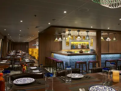 Innenansicht des Los Lobos Spezialitätenrestaurants an Bord der Norwegian Dawn. Lange Reihe gedeckter Tische, dahinter eine helle Bar mit blauen Kacheln und Barhockern. Im Hintergrund gelbe Sitznischen.