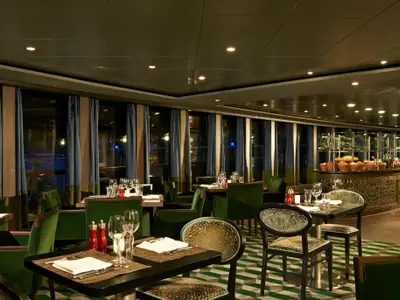 Spezialitätenrestaurant Moderno an Bord der Norwegian Dawn. Elegante Tische mit grünen Stühlen, Buffet und Fensterfront.