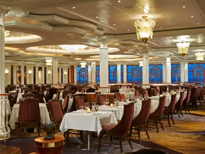 Das elegante Venetian Restaurant an Bord der Norwegian Dawn. Grosser Speisesaal mit weiss gedeckten Tischen, roten Stuehlen, Saeulen, Kronleuchtern und runden Fenstern mit Meerblick.