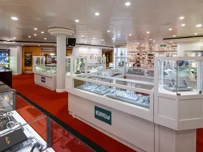 Ein heller Schmuckladen an Bord der Norwegian Dawn mit mehreren beleuchteten Vitrinen, die Schmuckstücke und Uhren präsentieren. Der Boden ist mit einem leuchtend roten Teppich ausgelegt. Im Hintergrund sind weitere Geschäfte erkennbar.