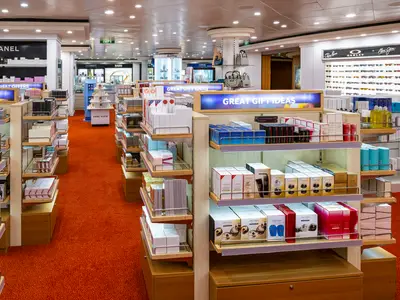 Ein Duty-Free-Shop an Bord der Norwegian Dawn. Der Gang mit orangefarbenem Teppich ist gesäumt von Regalen mit Kosmetik, Parfüm und Sonnenbrillen. Schilder 'GREAT GIFT IDEAS'.