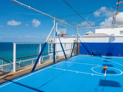 Ein leuchtend blauer Sportplatz an Bord der Norwegian Dawn, umgeben von einem Sicherheitsnetz, mit Basketballkorb, einem Fussballtor und einem orangen Basketball. Im Hintergrund ist das blaue Meer und der Himmel sichtbar.