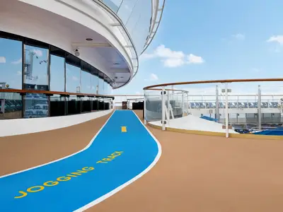Der blaue Jogging-Track mit gelber Markierung auf dem Oberdeck der Norwegian Dawn. Das braune Deck ist von Glaswänden und Geländern gesäumt. Im Hintergrund Liegestühle und blauer Himmel.