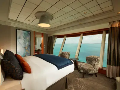 Geräumige, moderne Suite an Bord der Norwegian Dawn mit großem Bett und Blick auf das Meer. Große Fensterfront, zwei Sessel und abstrakte Kunst zieren den eleganten Raum.