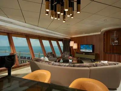 Luxuriöser Wohnbereich einer Suite an Bord der Norwegian Dawn. Mit beigem Rundsofa, schwarzem Flügel und großen Fenstern mit Meerblick.
