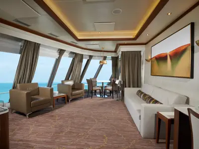 Blick in die helle und moderne Deluxe Owner's Suite an Bord der Norwegian Dawn mit braunen Ledersesseln, weißem Sofa und Esstisch, umgeben von großen Panoramafenstern mit Meerblick. Ein abstraktes Kunstwerk schmückt die Wand.