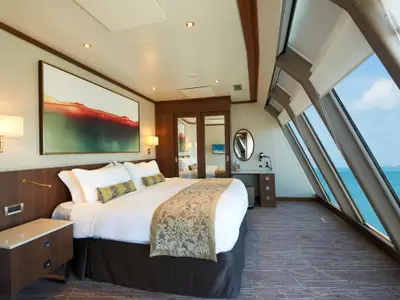 Eine helle, luxurioese Deluxe Owners Suite an Bord der Norwegian Dawn mit grossem Doppelbett, einem Beistelltisch, einem Arbeitsbereich und schraegen Fenstern, die einen weiten Meerblick bieten.