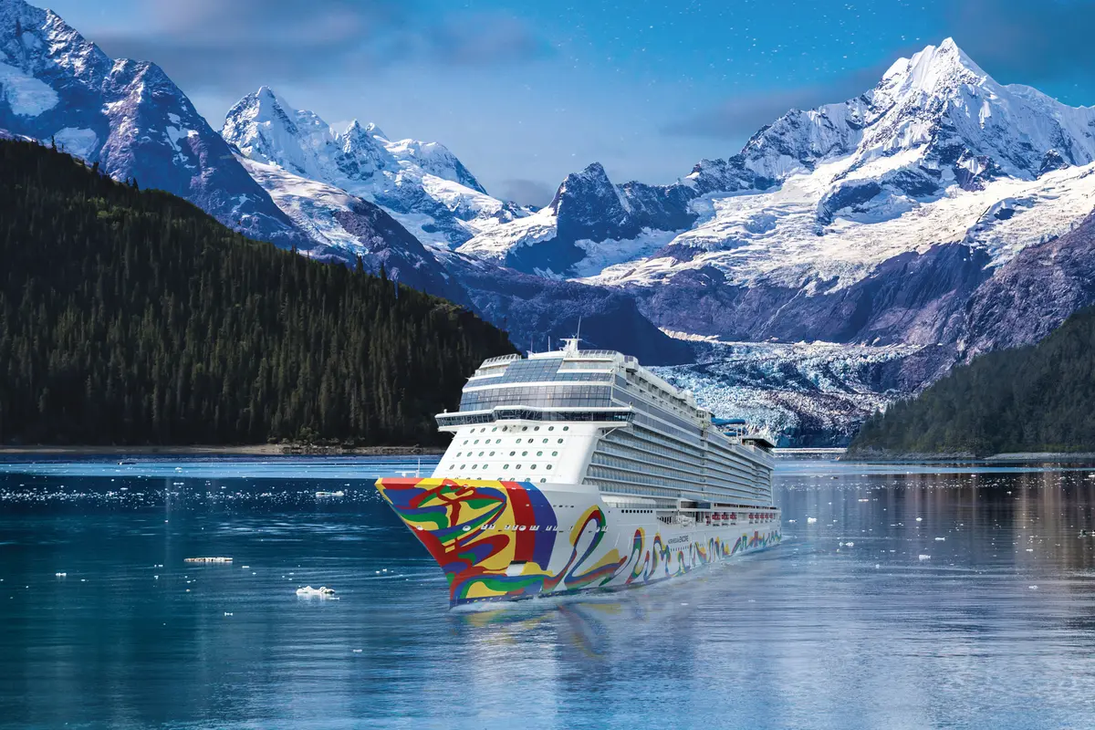 Die Norwegian Encore fährt mit farbenfrohem Bug durch einen Fjord in Alaska. Schneebedeckte Berge, Gletscher und viel Treibeis umgeben die Norwegian Encore unter blauem Himmel.