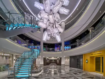 Das moderne Atrium der Norwegian Encore zeigt einen großen Kronleuchter, blau leuchtende Glastreppen, ein Casino und den gelben Social Club. Helle, elegante Innenarchitektur ohne Personen.