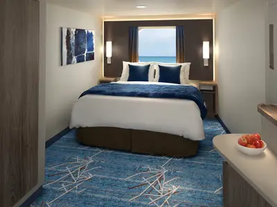 Moderne Aussenkabine auf der Norwegian Encore. Grosses Bett mit blauen Kissen und Decke. Fenster mit Meerblick. Links Wandkunst, rechts Beistelltisch mit Äpfeln.