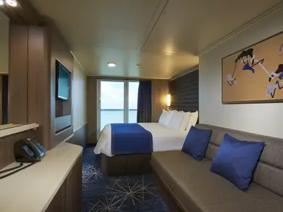 Helle Balkonkabine an Bord der Norwegian Encore mit großem Doppelbett, blauem Überwurf und gemütlichem Sofa mit blauen Kissen. Ein Balkon bietet Meerblick.