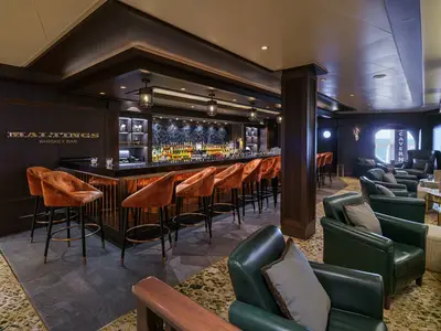 Die Maltings Whiskey Bar an Bord der Norwegian Encore zeigt eine lange Bar, Samt-Barhocker und grüne Ledersessel, mit Meerblick durch ein Fenster.