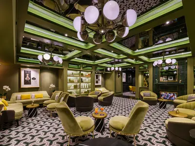 Eine luxuriöse, leere Bar-Lounge an Bord der Norwegian Encore mit grünen Polstermöbeln, gemustertem Teppich und einer auffälligen Deckenbeleuchtung, inklusive Drinks auf einem Tisch.