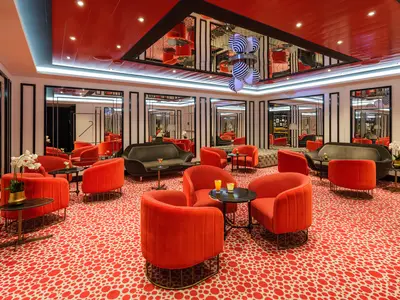 Moderne Bar-Lounge an Bord der Norwegian Encore. Rote Sessel und Sofas, gemusterter Teppich. Spiegelwände und eine Deckenleuchte mit schwarz-weißen Kugeln. Getränke auf Tischen.
