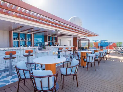 Die helle Spice H2O Bar auf dem Sonnendeck der Norwegian Encore zeigt Barhocker, Tische mit Stühlen und Champagnergläser. Im Hintergrund ist bunte Wandkunst zu sehen.