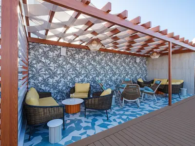 Überdachte Outdoor-Lounge auf der Norwegian Encore mit Palmen-Tapete. Blaue Mosaikfliesen und moderne Rattanmöbel in Gelb und Blau unter einem Holz-Pergola-Dach.