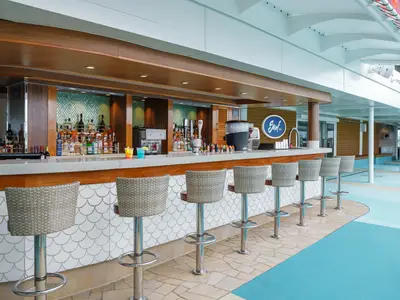 Blick auf die leere Surf Bar an Bord der Norwegian Encore. Eine lange Theke mit vielen Flaschen, Hocker und bunte Drinks sind zu sehen. Die Rückwand ist mit türkisen Mosaikfliesen und einer runden Bar-Plakette versehen.