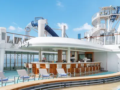 Die Waves Pool Bar auf der Norwegian Encore: Bartheke mit Hockern und Sonnenliegen. Im Hintergrund: Wasserrutschen, Wendeltreppe und das Meer unter blauem Himmel.
