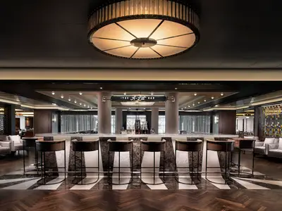 Innenansicht der luxuriösen A-List Bar an Bord der Norwegian Encore mit einer langen Bar, Barhockern, Marmorverkleidung und einer beeindruckenden runden Deckenlampe. Sitzbereiche befinden sich seitlich, der Boden ist gemustert.