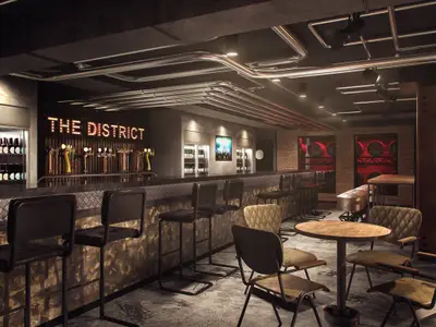 Blick in die moderne Bar "The District" an Bord der Norwegian Encore. Zu sehen sind eine lange Theke, Barhocker, Zapfanlagen, Flaschenkühlschränke und ein leuchtender Schriftzug. Die Beleuchtung ist gedämpft und schafft eine stimmungsvolle Atmosphäre.