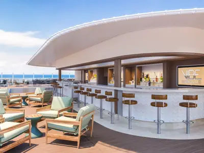 Rendering des Vibe Beach Club an Bord der Norwegian Encore. Eine moderne Outdoor-Bar mit Barhockern und Loungesesseln auf dem Deck. Im Hintergrund ist das Meer unter blauem Himmel zu sehen.